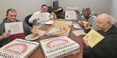 Four men eating pizza and holding up certificates