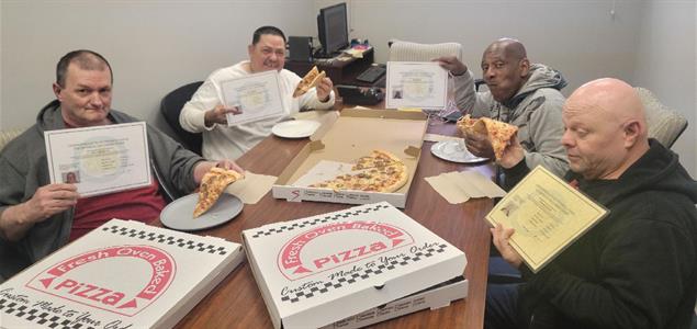 Four men eating pizza and holding up certificates