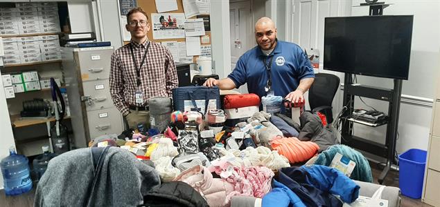 Two people stand behind a table full of donated clothes and blankets.