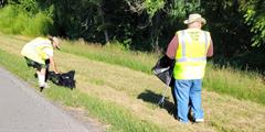 Two men pick up trash along the side of the road