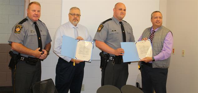 Two men stand with two PA State Troopers holding certificates
