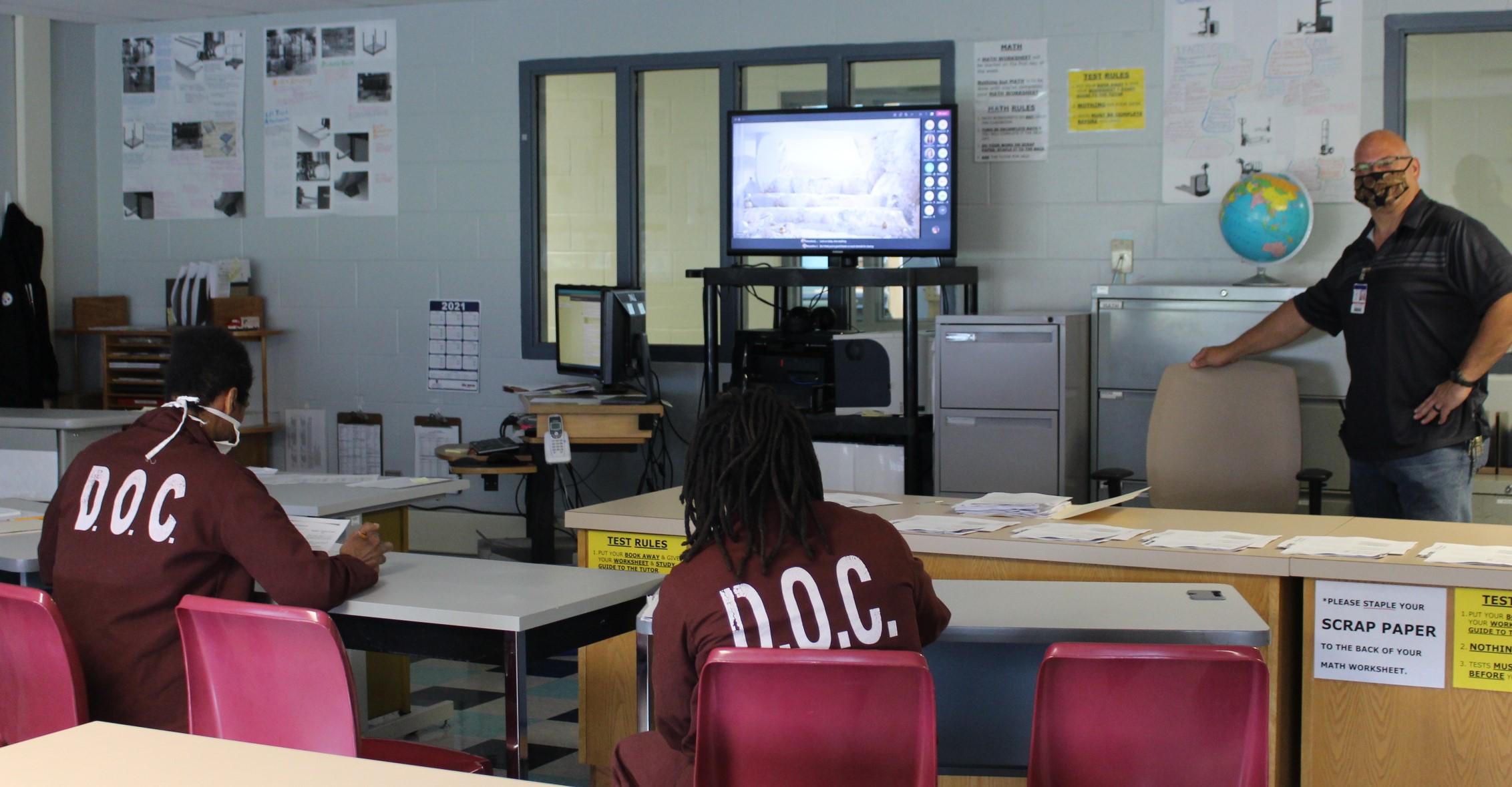 Two inmates watch a virtual presentation with an employee standing by observing