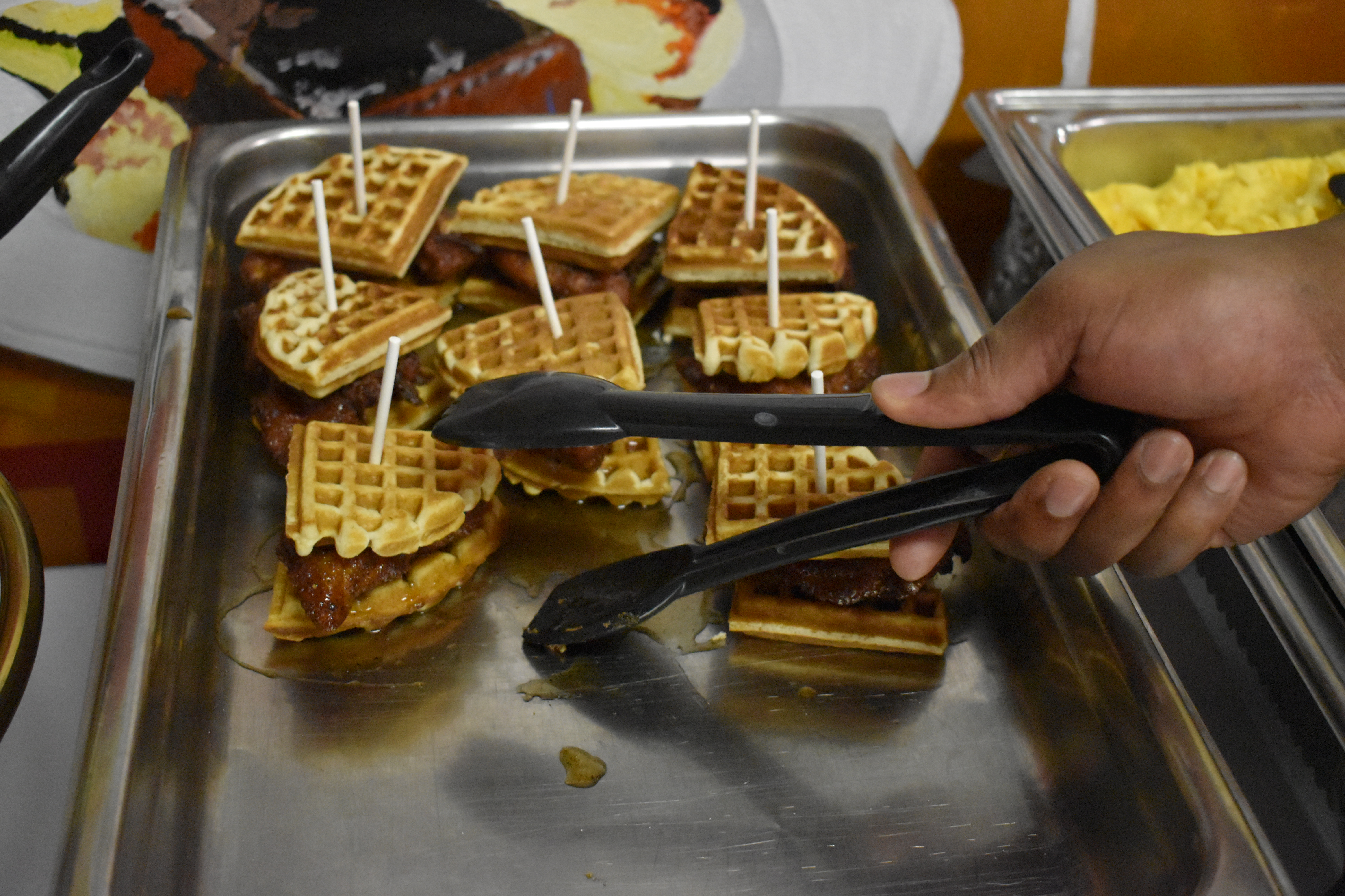 A hand uses a utensil to grab a chicken and waffle slider from a tray