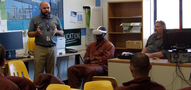 An inmate sits while using virtual reality goggles as an employee explains the program to other inmates.