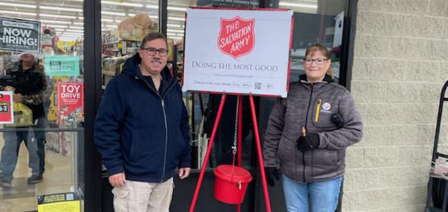 Two people stand by a Salvation Army kettle to ring a bell.