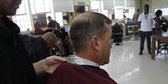 A barber wraps a sheet around a man sitting for a haircut while others look on