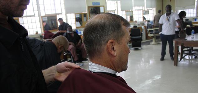 A barber wraps a sheet around a man sitting for a haircut while others look on