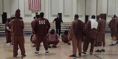 A group of incarcerated men listen to a guest speaker in a gym