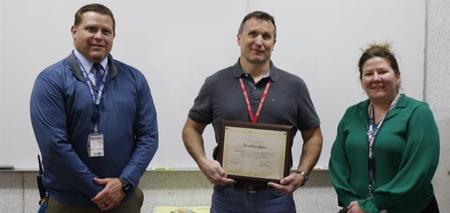 Psychological Service Specialist Thaddeus Gross stands between two people while he holds a certificate.
