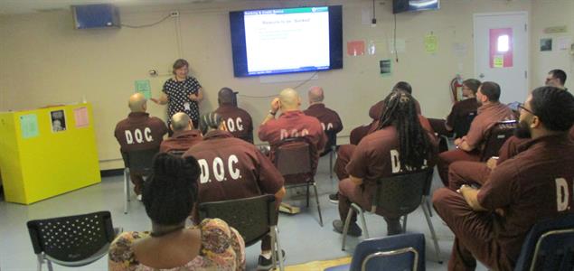 A group of inmates sit watching a speaker at the front of a classroom