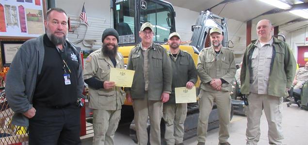 Six people stand in front of a construction vehicle as two hold certificates.
