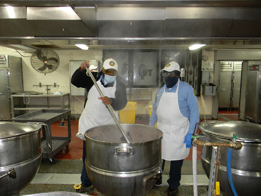 Two food service employees mix a vat in the kitchen