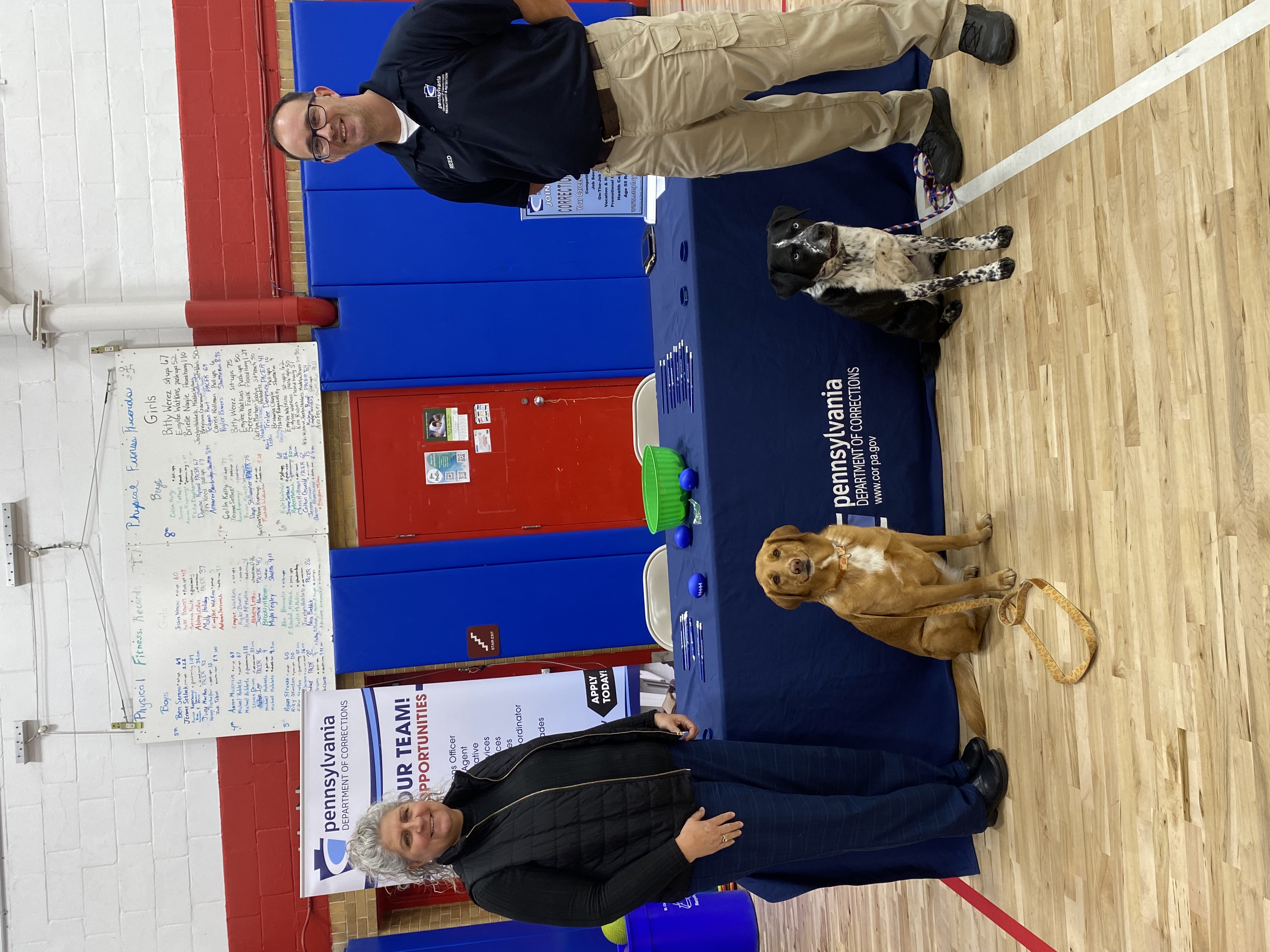 Lt. Reed, Unit Manager Marhelko and two dogs stand in front of a table at a local career fair.